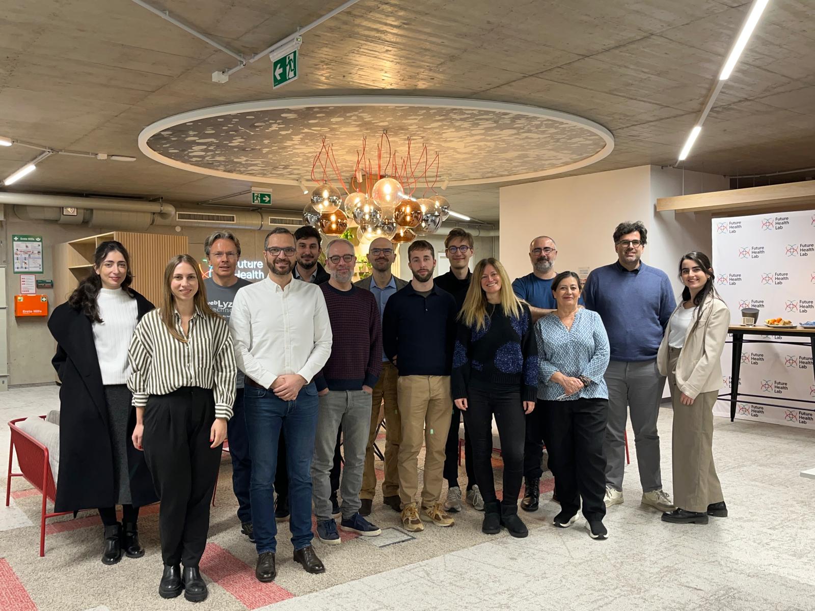 A group of about fifteen people pose together indoors beneath a decorative round ceiling fixture with hanging light bulbs, with a "Future Health Lab" backdrop visible on the right.