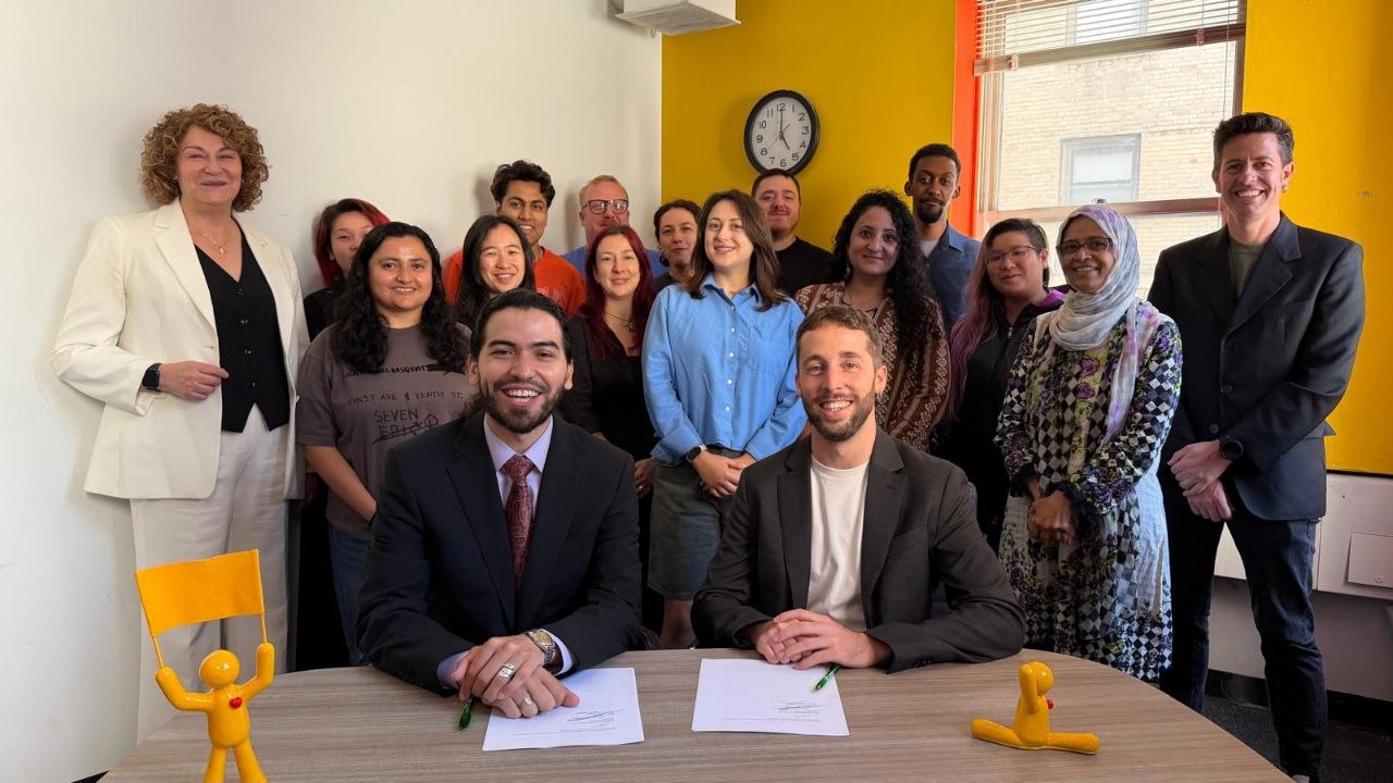 Two men in suits sit at a table with signed documents in front of them, smiling alongside a larger group of people standing behind them in an office with yellow walls.